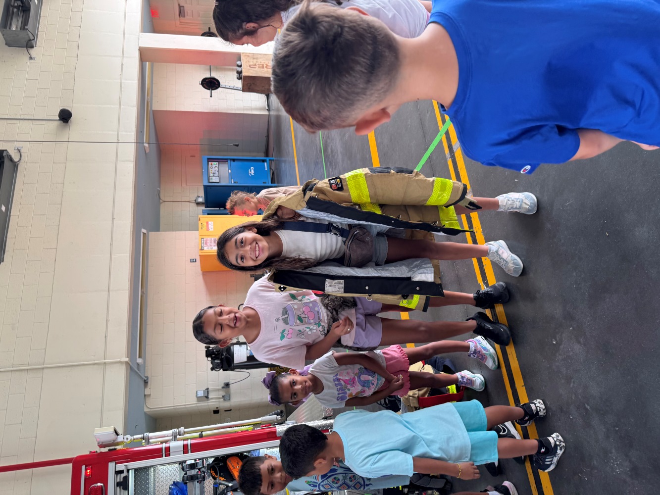 Children gather around a girl wearing an oversized firefighter coat inside a fire station bay